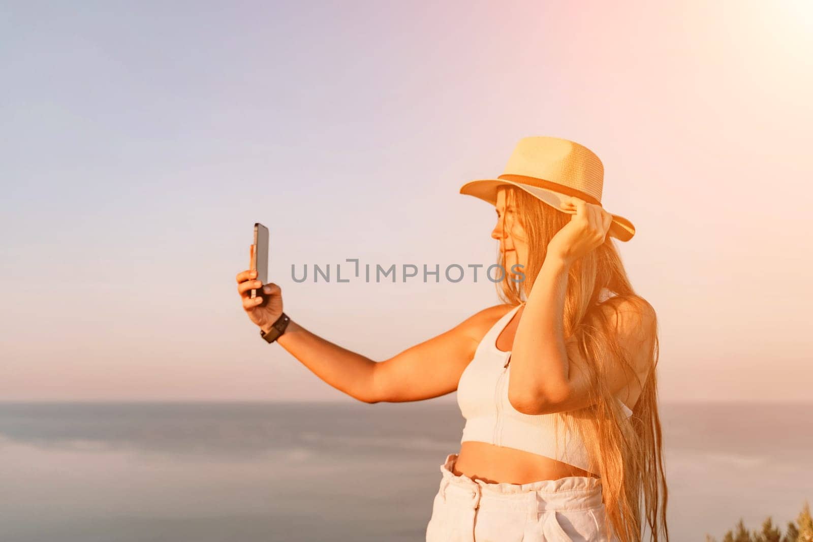 Selfie woman in a hat, white tank top, and shorts captures a selfie shot with her mobile phone against the backdrop of a serene beach and blue sea