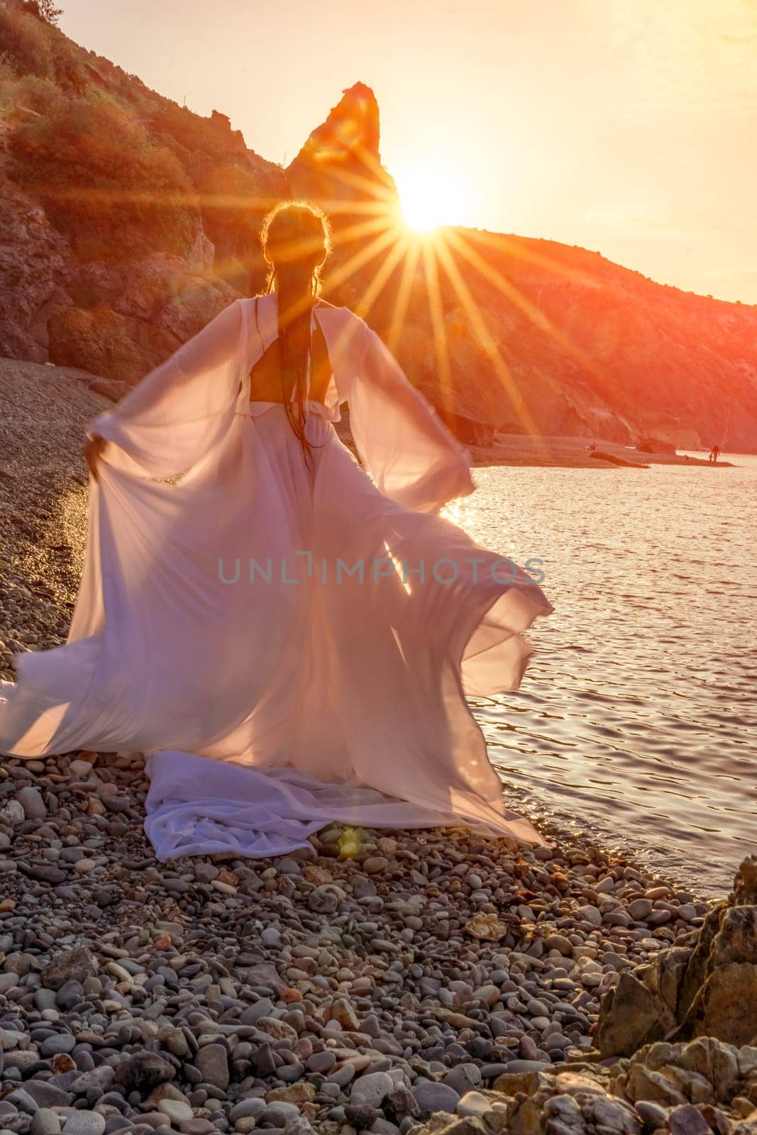 Mysterious woman silhouette long hair walks on the beach ocean water, sea nymph wind listens to the wave. Throws up a long white dress, a divine sunset. Artistic photo from the back without a face.