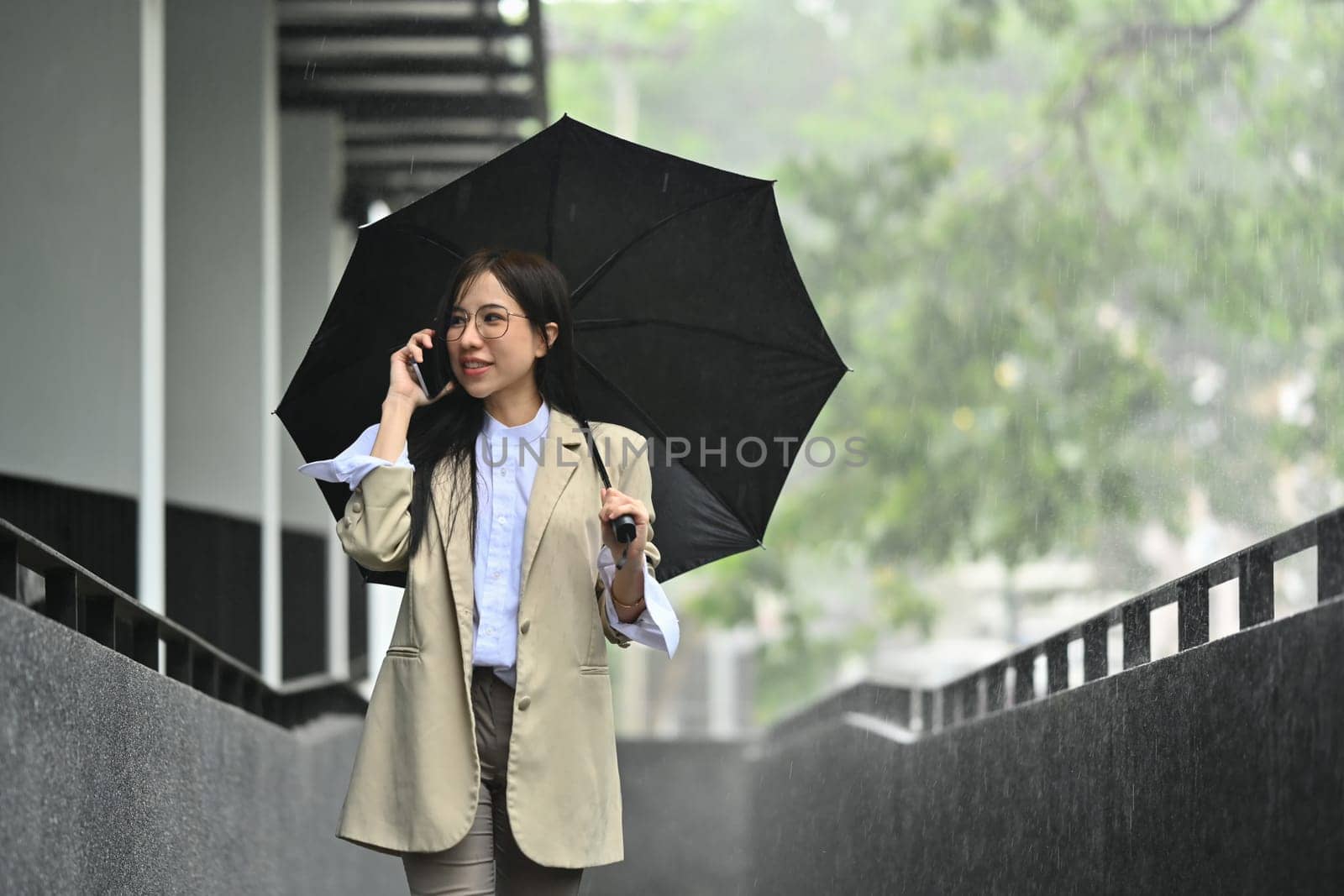 Gorgeous millennial businesswoman talking on mobile phone under umbrella while walking in the city on rainy day.