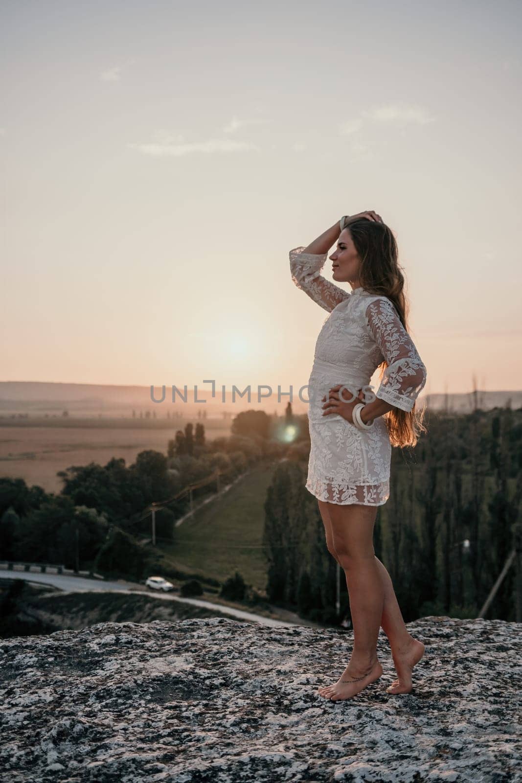 Happy woman in white boho dress on sunset in mountains. Romantic woman with long hair standing with her back on the sunset in nature in summer with open hands. Silhouette. Nature. Sunset. by panophotograph