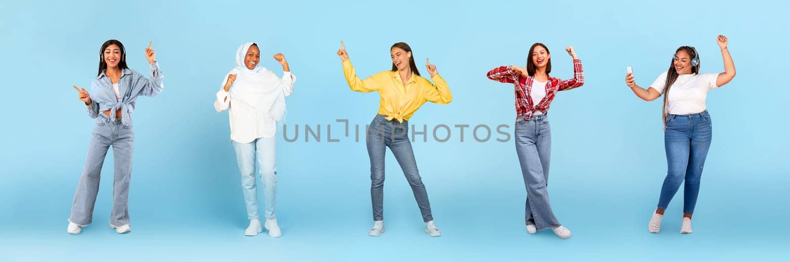 Smiling millennial diverse women in wireless headphones dancing, have fun with phone, enjoy fun, isolated on blue studio background, panorama, full length. Relax, lifestyle, ad and offer