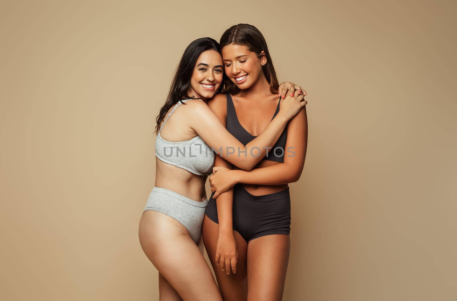 Cheerful millennial latin, arab women in underwear hugs, isolated on beige background, studio. Motivation for self love, spa treatments and fit, wellness, skin care, natural beauty