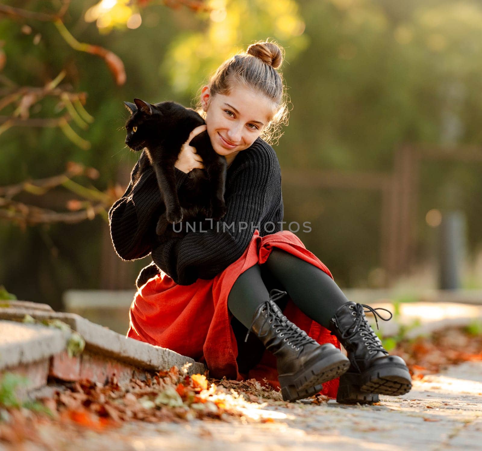 Pretty girl in red skirt hugging black cat outdoors at street with autumn leaves. Beautiful model teenager sitting with feline animal at park