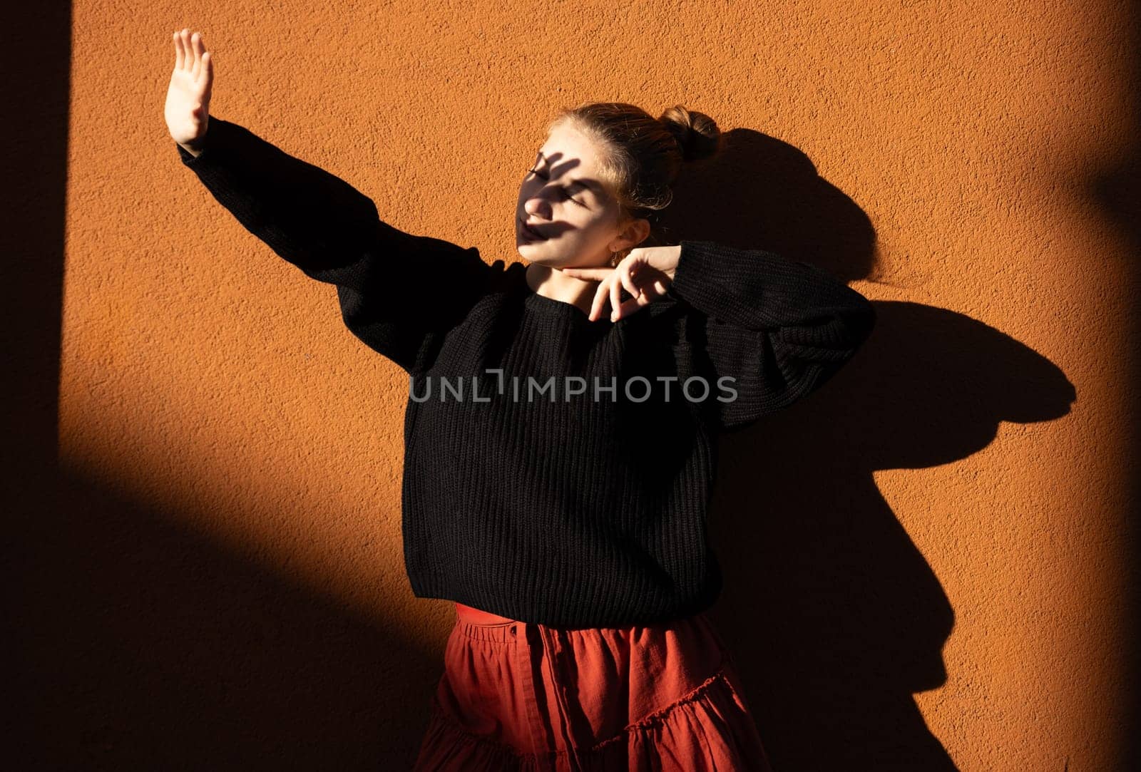 Pretty girl posing near wall with shadows covering face with hand from sunlight. Beautiful teenager model in sun rays at autumn
