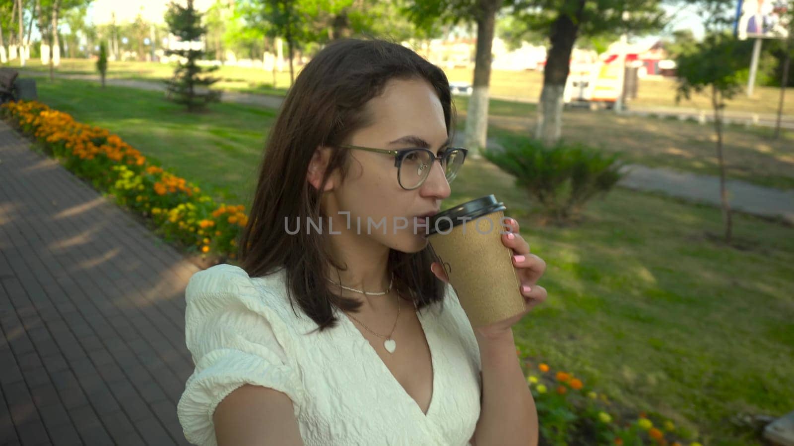 A young woman walks along an alley in a white dress and drinks crofe. A girl with glasses walks through the park and films herself on camera. 4k