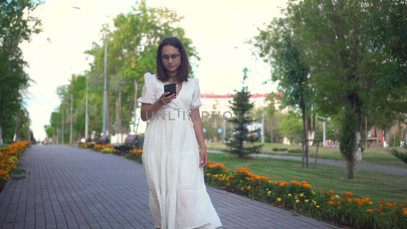 A young woman walks along a flower alley with a phone in her hands. A girl in a white dress looks at her phone and walks among the flowers. 4k