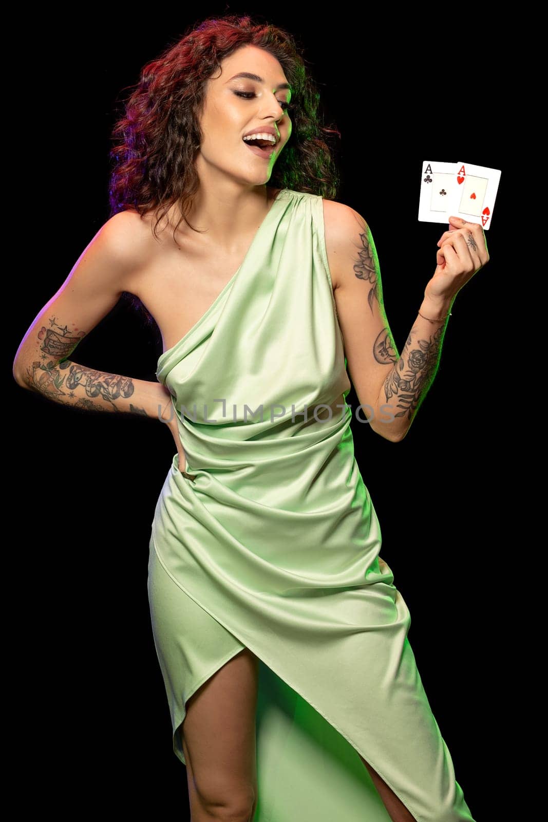 Cheerful young woman with wavy dark hair and tattoos on arms wearing light green dress standing against black background, holding set of winning cards of pair of aces. Successful gambling concept