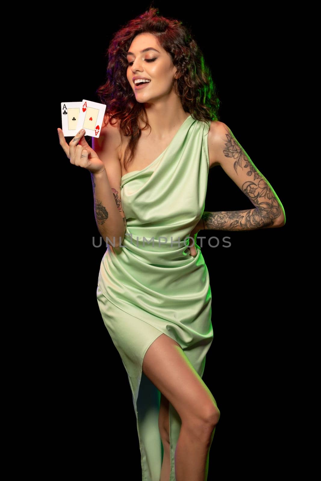 Portrait of cheerful young brunette with wavy hair wearing elegant light green dress, holding two aces in hands, posing against black background. Poker win concept