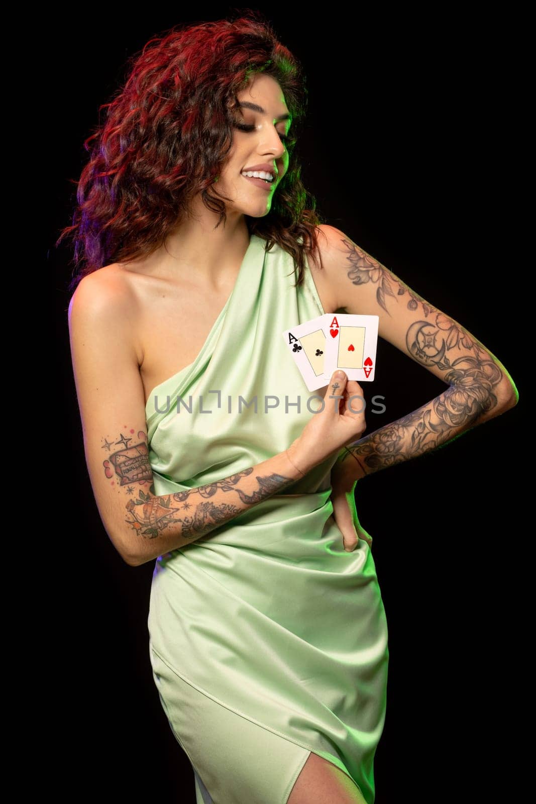 Successful young female poker player in light green dress with naked shoulder and tattoo on arms standing against black background, holding winning pair of aces. Gambling concept