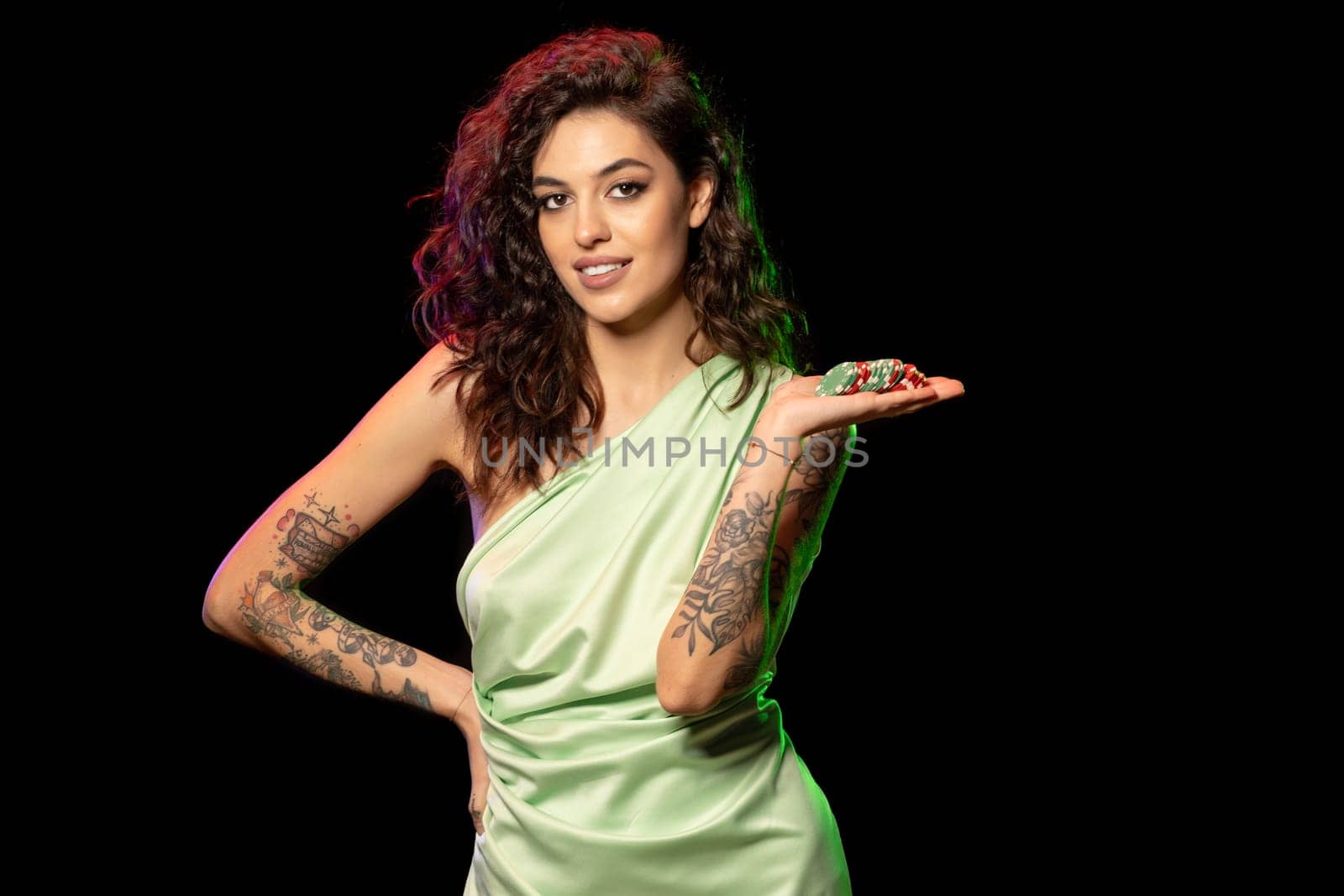 Confident happy young female gambler, avid poker player, holding pile of multicolored betting chips in palm standing on dark studio background. Concept of passion for gambling and winning