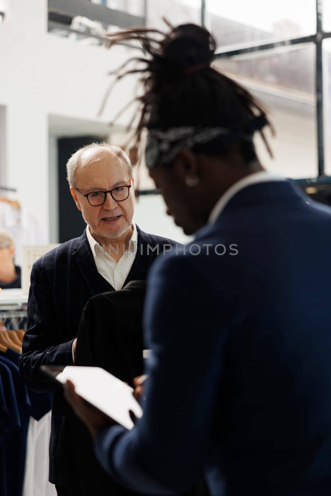 Elderly customer asking employee to check shirt inventory on tablet computer, wanting to buy formal wear in clothing store. Diverse people discussing clothes fabric in modern boutique