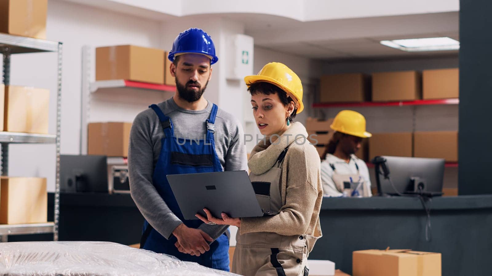 Diverse employees talking about stock logistics in depot, merchandise in boxes placed on storage room racks. Man and woman wearing overalls and hardhats using laptop in warehouse space.