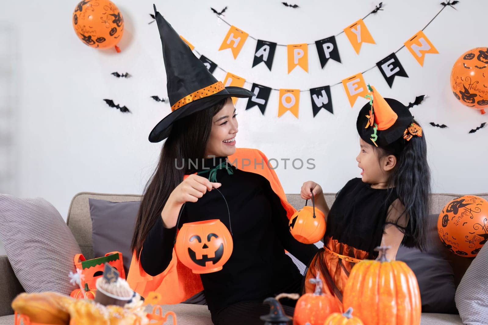 A playful young mother sits on a comfortable sofa in the living room decorated with pumpkins and her cute little daughter tells a spooky Halloween story. Funny mother and girl having fun at home by wichayada
