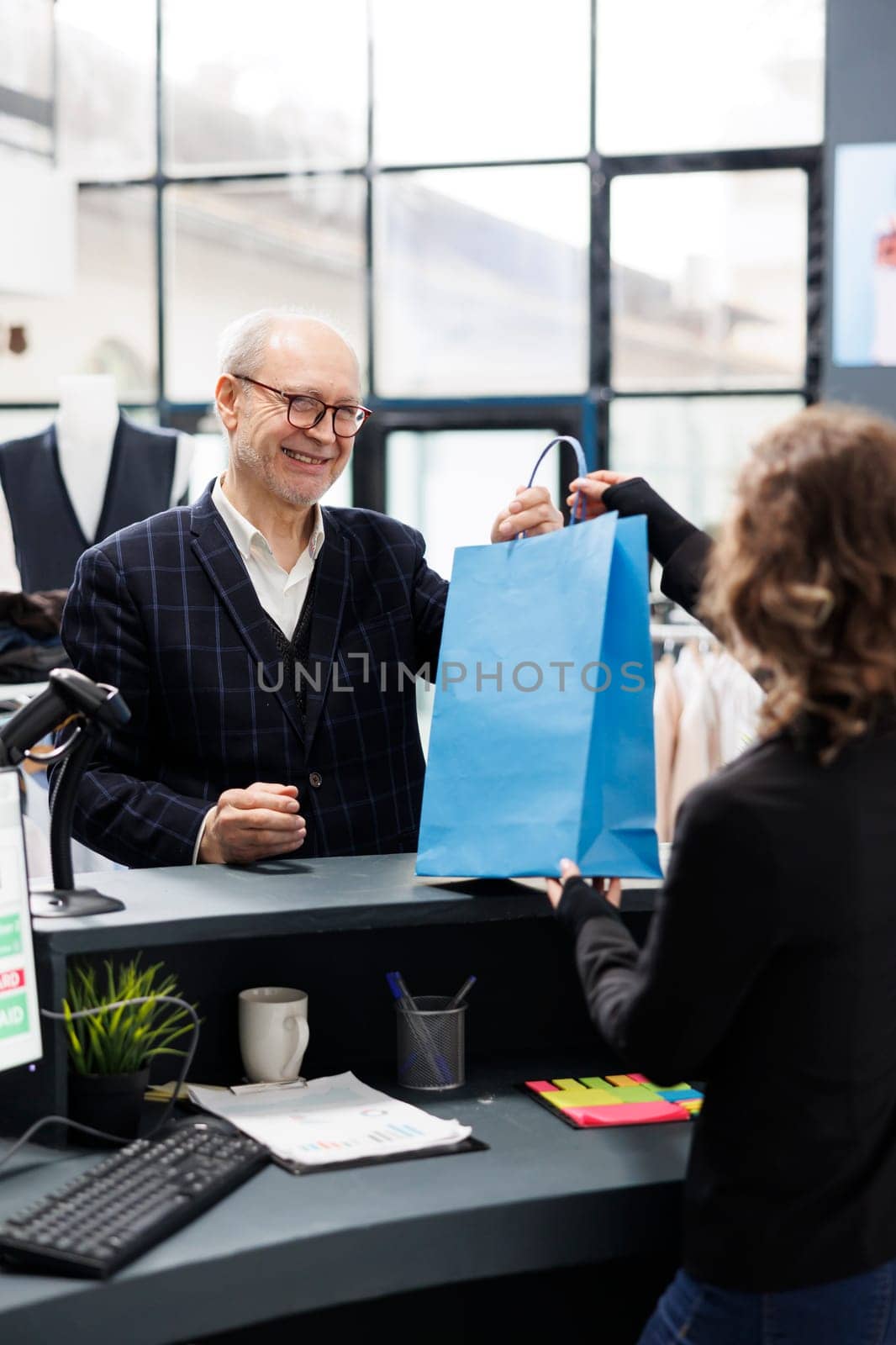 Senior client taking shopping bag from store worker, standing at counter desk paying for purchase in fashion boutique. Elderly man buying modern clothes making electronic transaction at mall