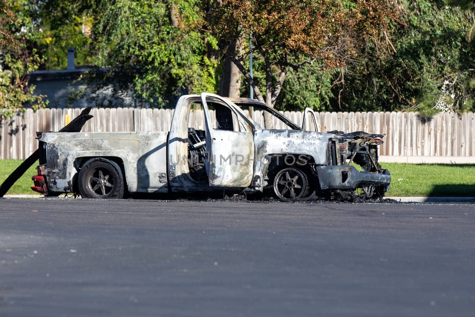 A burnt-out truck sits abandoned on a suburban street, its charred frame highlighting recent fire damage. Bright sunlight and greenery contrast with the vehicle's destruction, adding to the scene's stark realism.