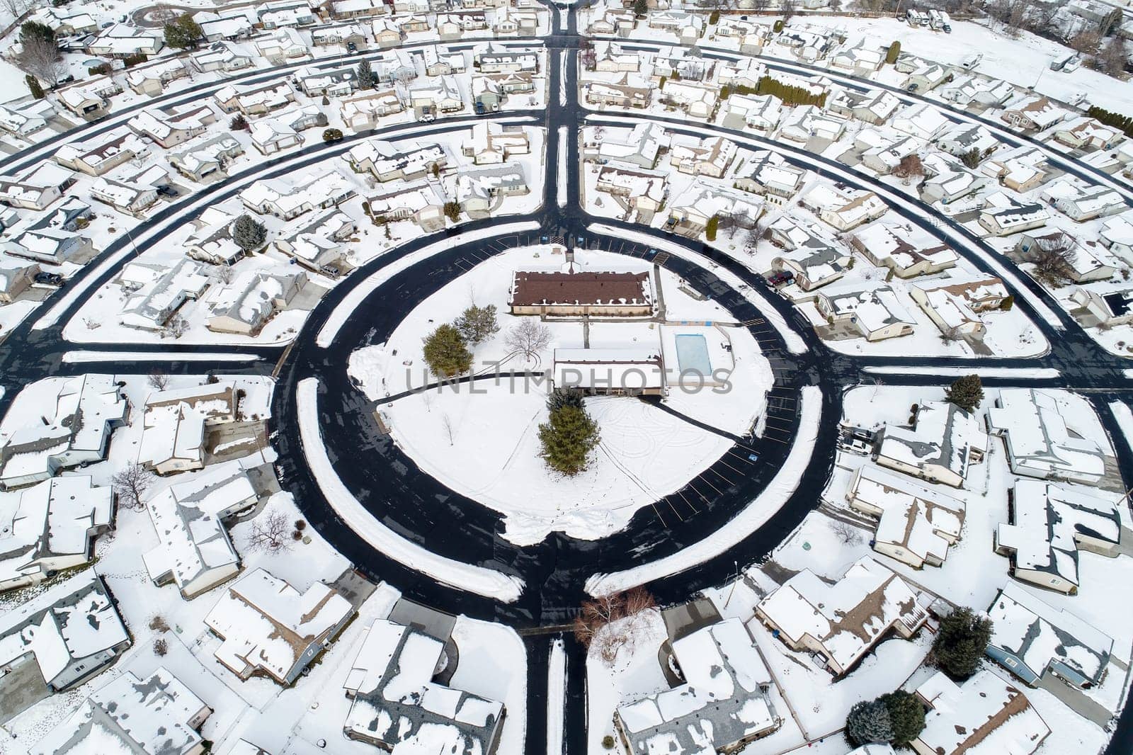 High-angle aerial photograph of a circular suburban neighborhood blanketed in snow, showcasing smooth roundabouts and rooftops. A peaceful winter scene captured from above.