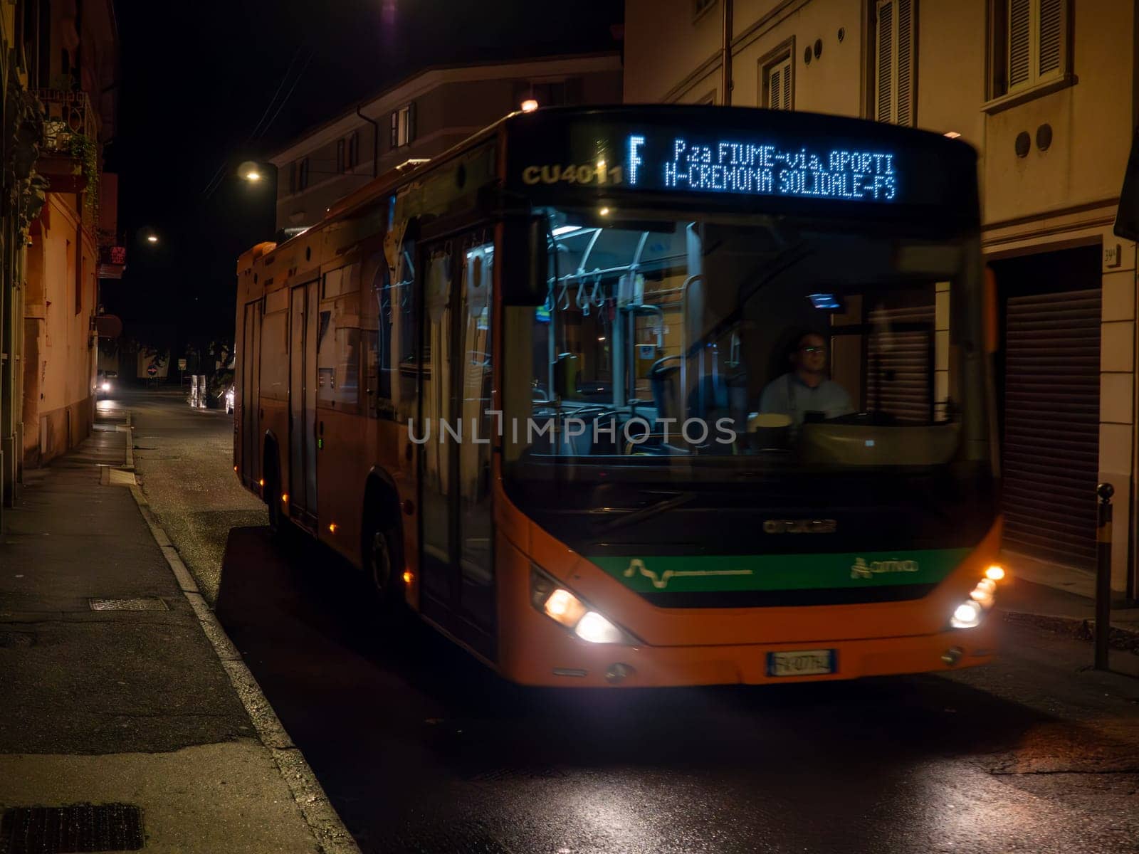 Cremona, Lombardy, Italy October 15th 2024 An electric bus is driving down a narrow, empty street at night, illuminating the wet asphalt with its headlights