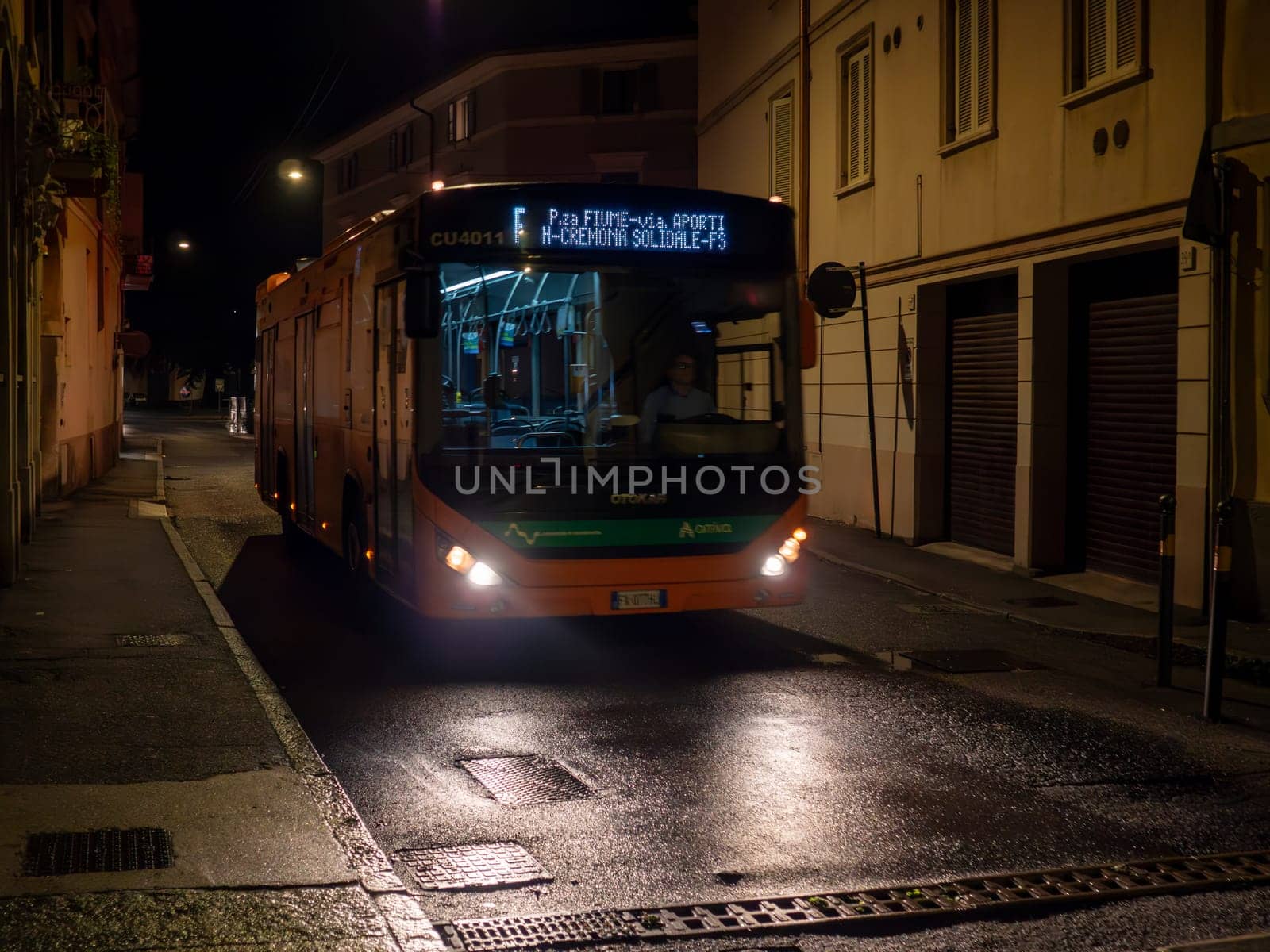 Cremona, Lombardy, Italy October 15th 2024 An electric bus is driving down a narrow, empty street at night, illuminating the wet asphalt with its headlights