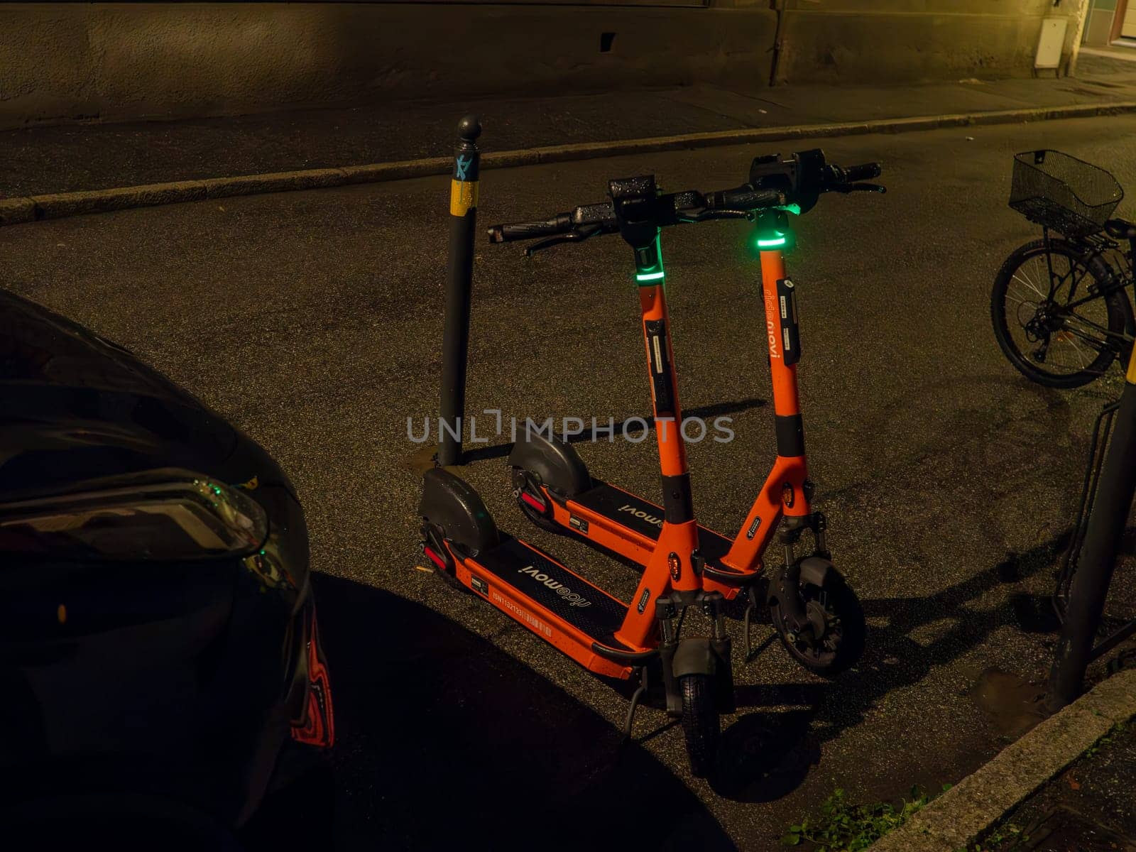 Cremona, Lombardy, Italy October 15th 2024 Two orange electric scooters parked next to a pole on a city sidewalk at night, illuminated by green lights