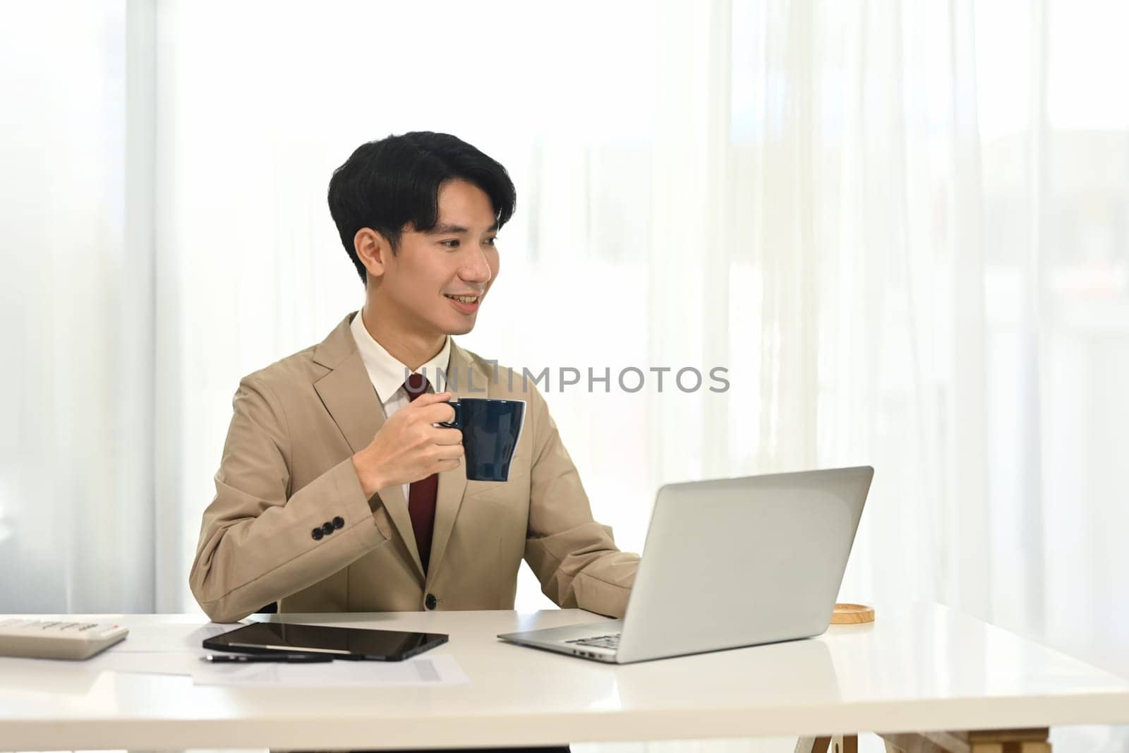 Young businessman drinking coffee and working with laptop in bright office.