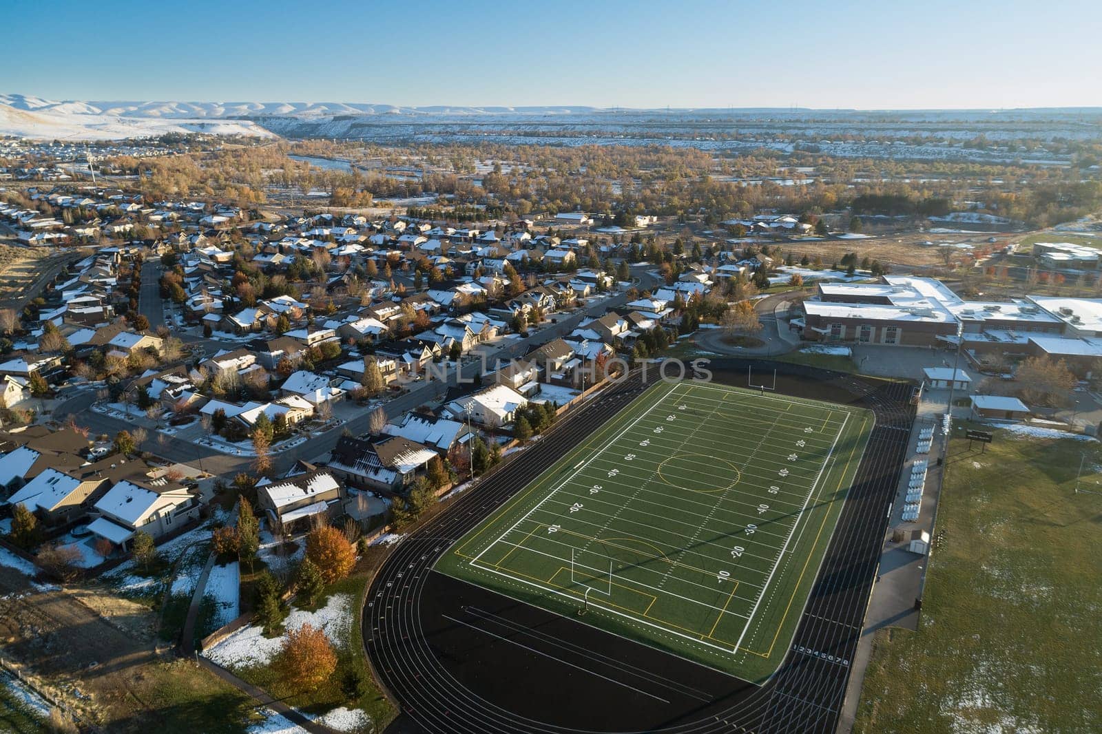 A stunning aerial photo of a suburban neighborhood featuring a football field surrounded by houses, trees, and distant hills. Captures the essence of community and outdoor recreation.
