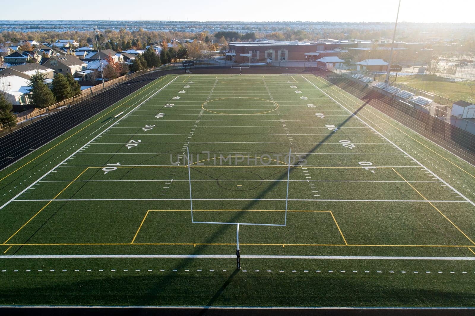 An expansive aerial view of a vacant football field surrounded by a serene neighborhood. The sun casts long shadows, adding depth to the vibrant green turf.