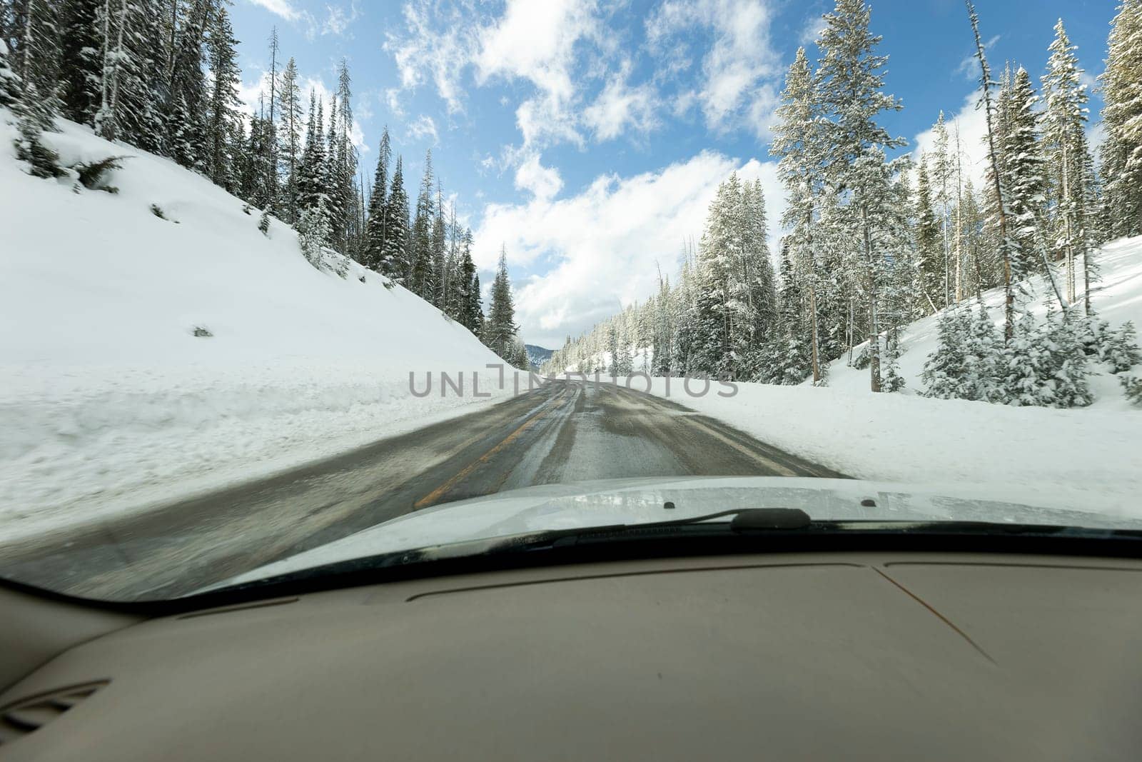 Inside view of a car driving through snow covered mountains in a forest with blue sky in the background