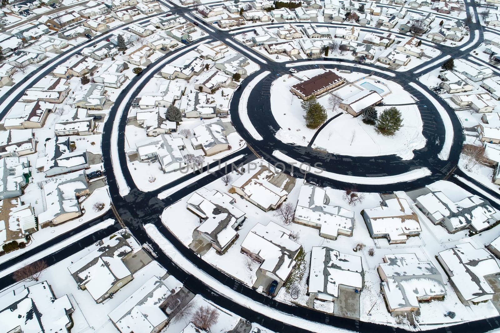 Aerial image showcasing a snow-blanketed suburban neighborhood. The circular road layout and uniform houses present a serene winter scene from above, highlighting the beauty of snowy landscapes.