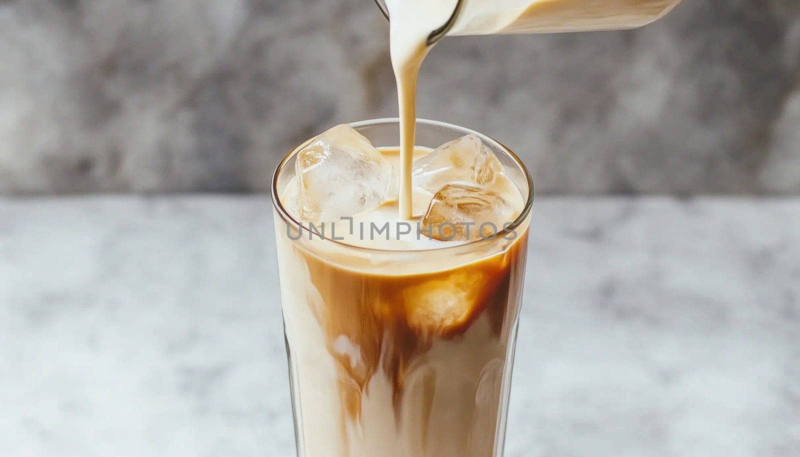 Milk being poured from a bottle into a glass of iced coffee with ice cubes on a marble countertop