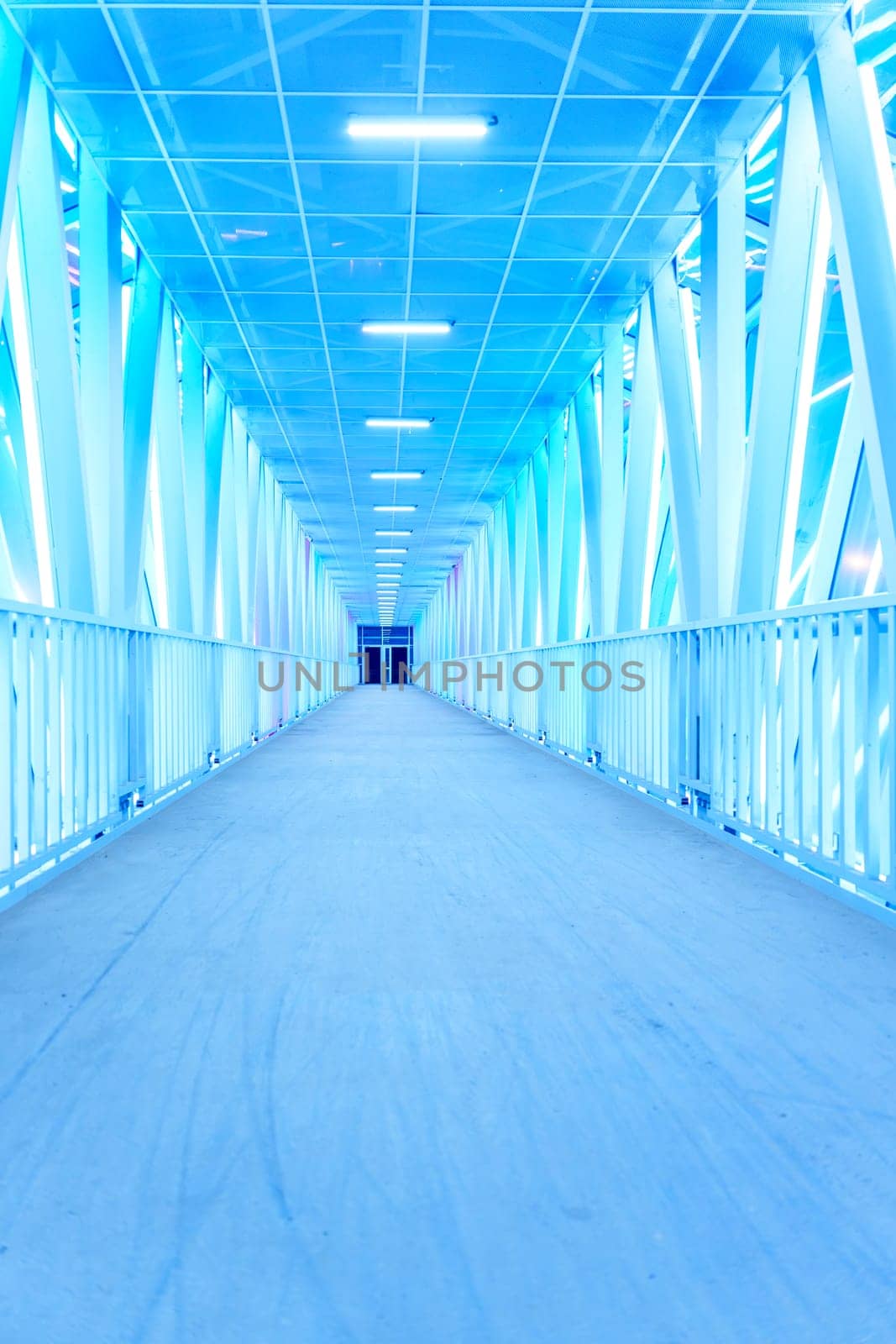 A futuristic illuminated walkway with a symmetrical design and glowing blue lights creates a modern, minimalist atmosphere. The empty corridor stretches into the distance with no people in sight