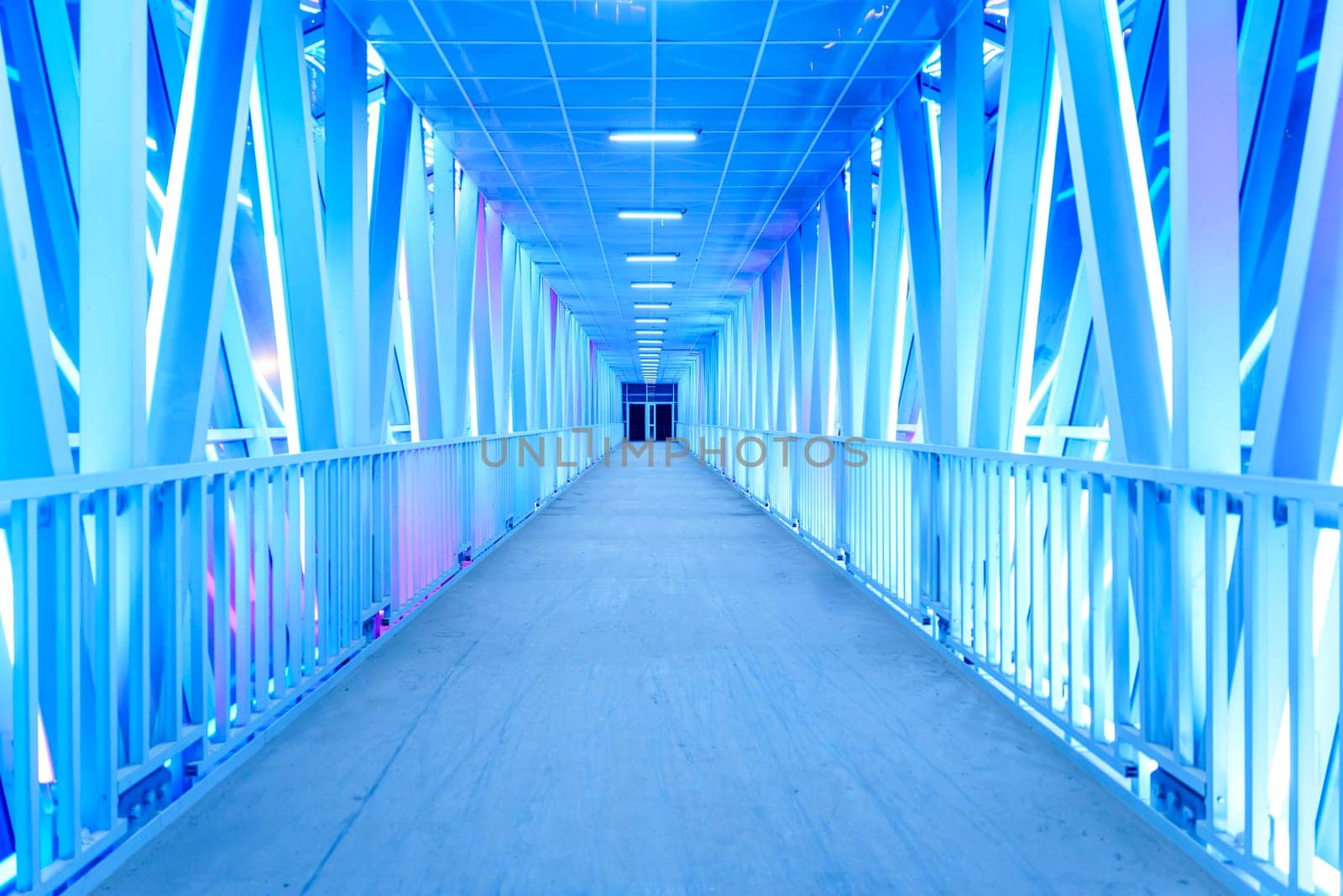A futuristic illuminated walkway with a symmetrical design and glowing blue lights creates a modern, minimalist atmosphere. The empty corridor stretches into the distance with no people in sight