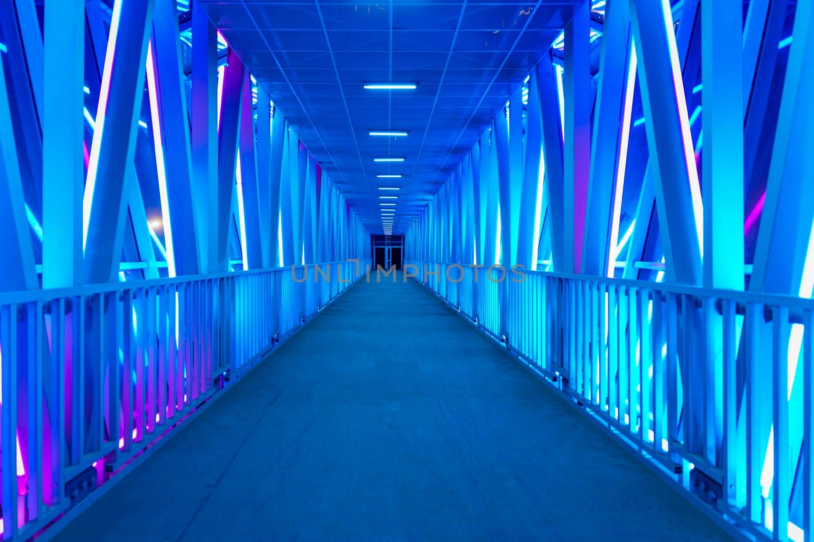 A futuristic illuminated walkway with a symmetrical design and glowing blue lights creates a modern, minimalist atmosphere. The empty corridor stretches into the distance with no people in sight