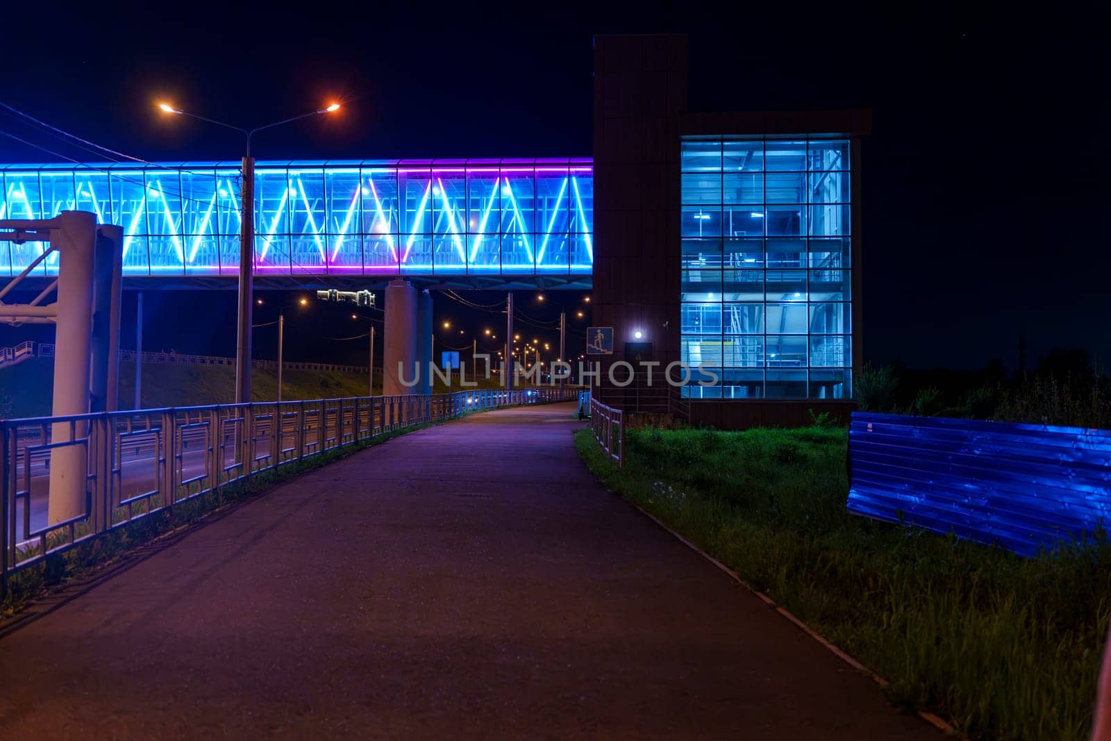 An illuminated pedestrian bridge with vibrant blue and purple lights connects to a modern glass building at night. The scene is peaceful and urban, with no people in sight