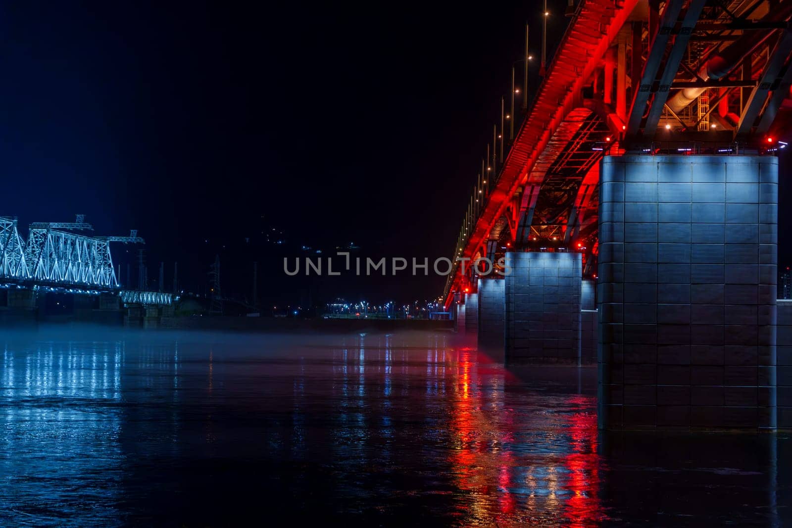 Red and blue illuminated bridges stretch over a river at night, with mist hovering over the water and reflections shimmering below. The serene urban scene captures the contrasting colors