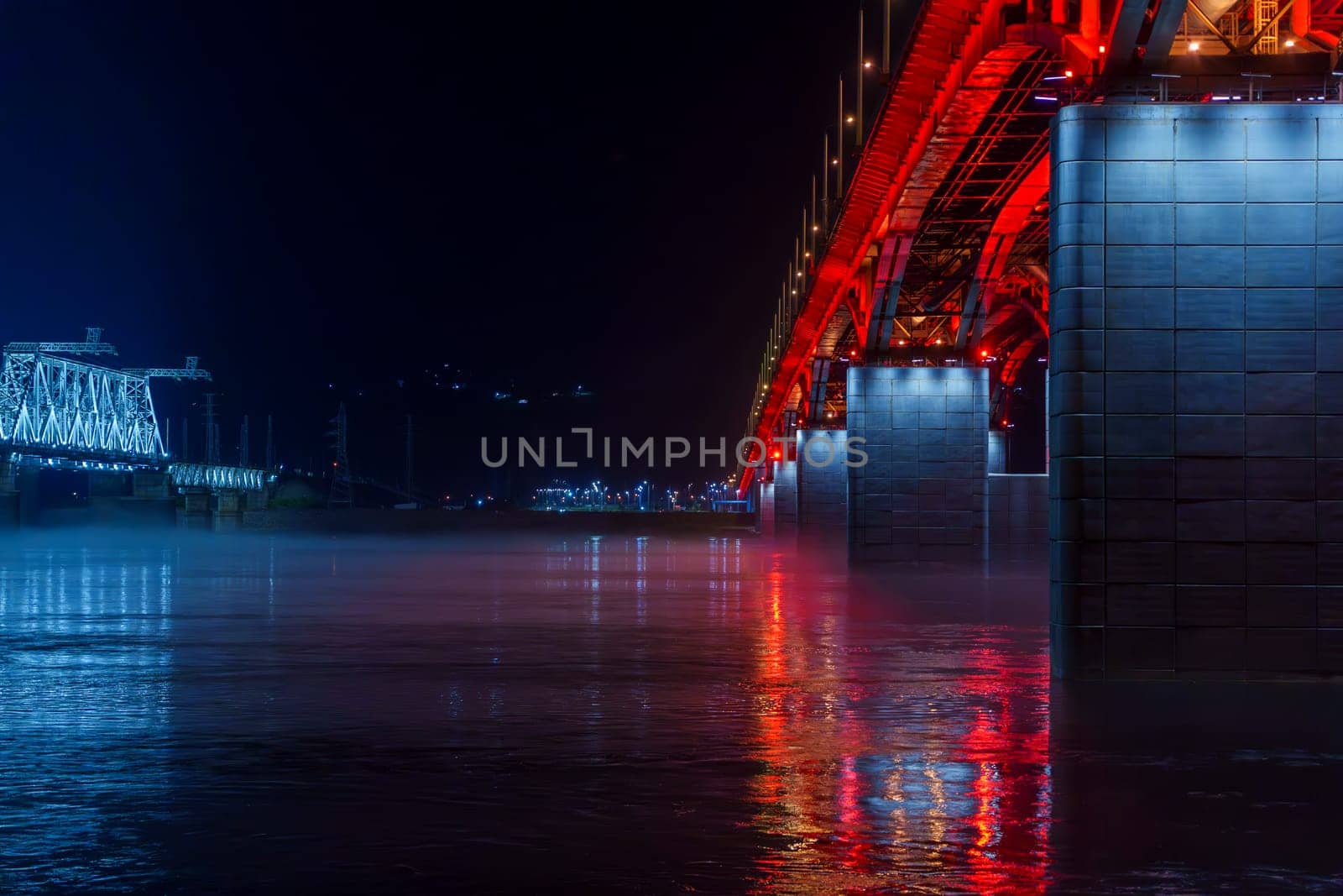 Red and blue illuminated bridges stretch over a river at night, with mist hovering over the water and reflections shimmering below. The serene urban scene captures the contrasting colors