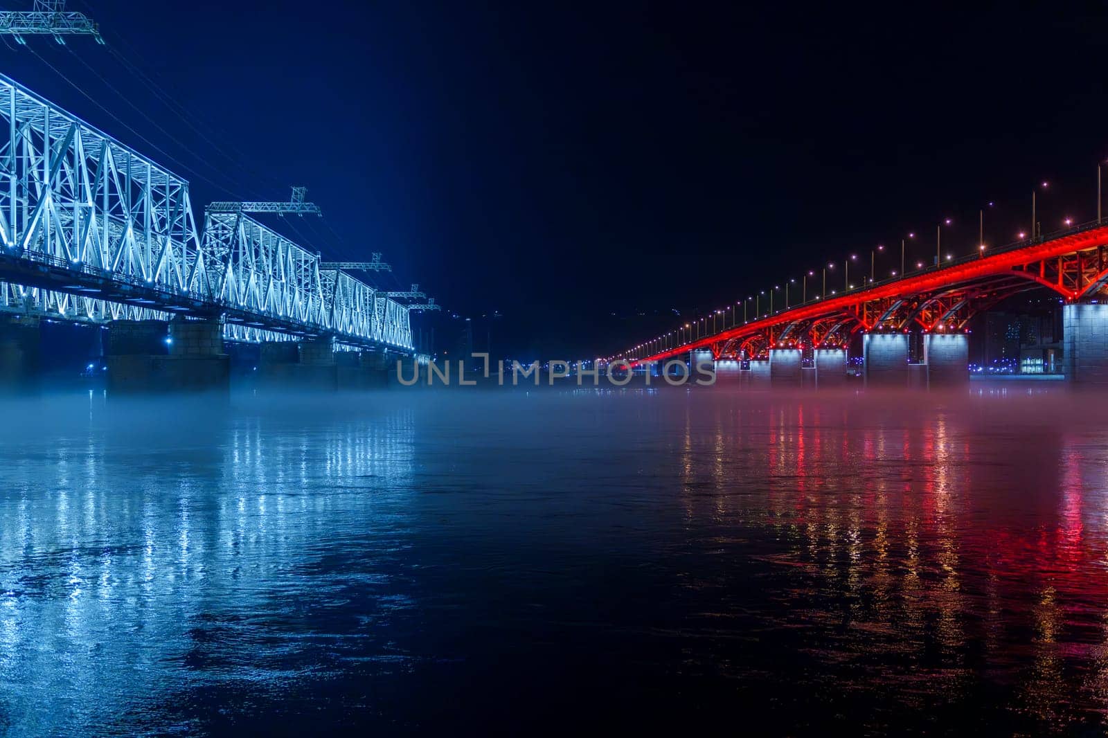 Two illuminated bridges, one blue and one red, stretch over a river at night. A layer of mist hovers over the water, reflecting the lights from the bridges