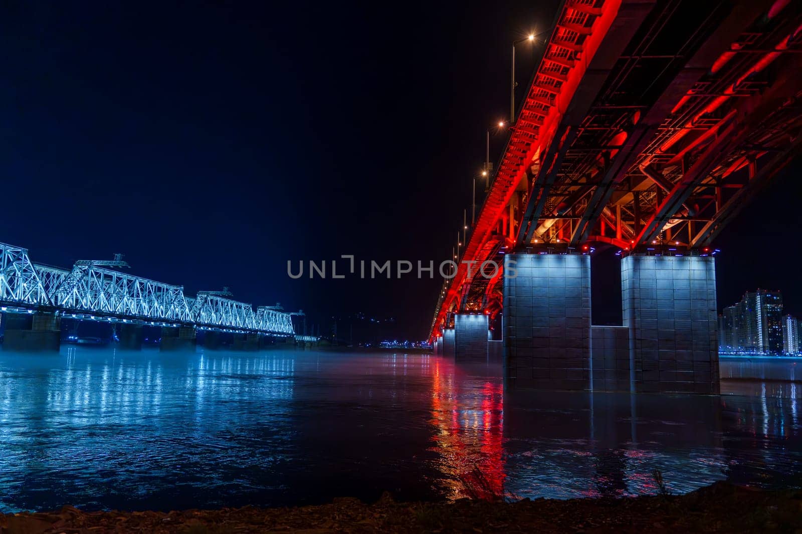 Red and blue illuminated bridges stretch over a river at night, with mist hovering over the water and reflections shimmering below. The serene urban scene captures the contrasting colors
