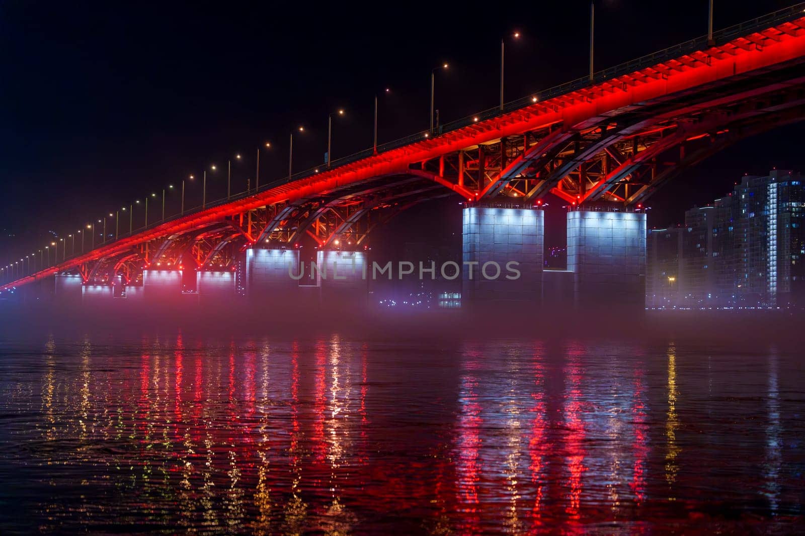 A red illuminated bridge stretches over a calm river at night, with mist floating above the water and colorful reflections. The serene urban scene captures the structure and lighting