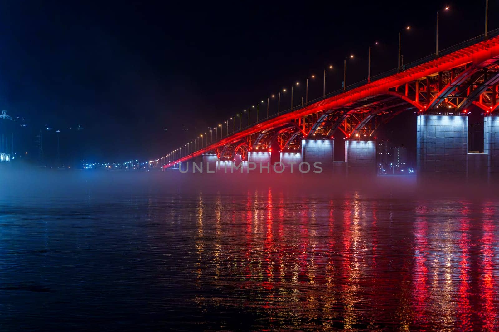 A red illuminated bridge stretches over a calm river at night, with mist floating above the water and colorful reflections. The serene urban scene captures the structure and lighting
