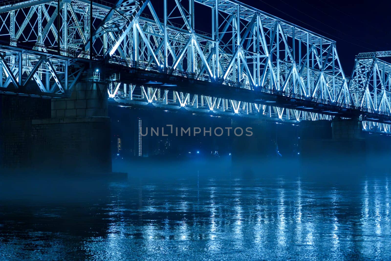 A blue illuminated steel bridge spans over a calm river at night, with mist hovering above the water and reflections shimmering below. The peaceful and serene urban scene shows no people present