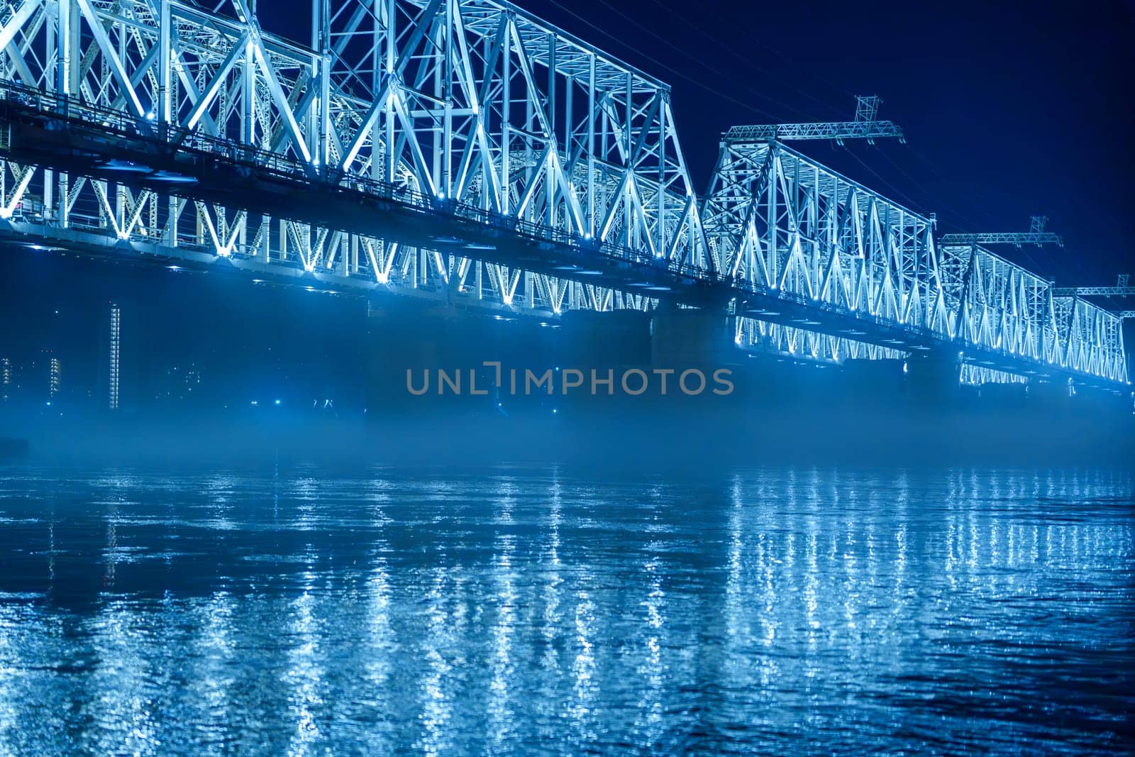 A blue illuminated steel bridge spans over a calm river at night, with mist hovering above the water and reflections shimmering below. The peaceful and serene urban scene shows no people present