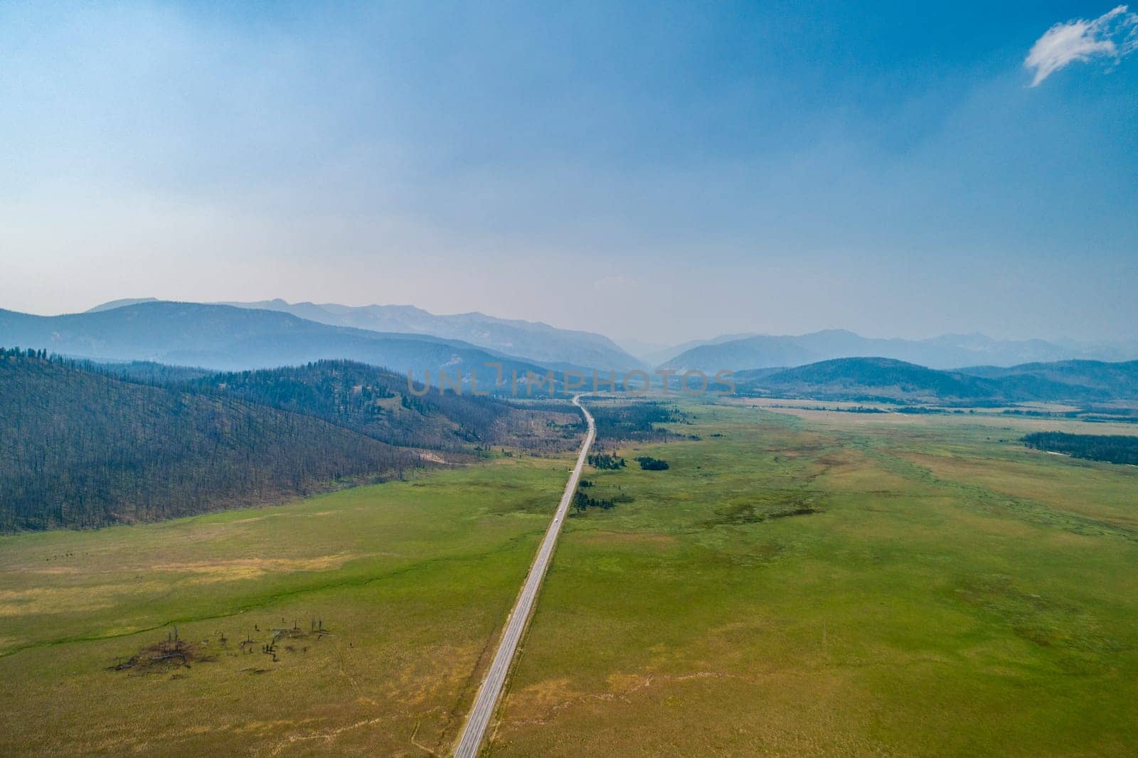 A long road stretches through expansive green plains towards distant mountains under a clear blue sky. The serene landscape offers a sense of peace and endless possibilities.