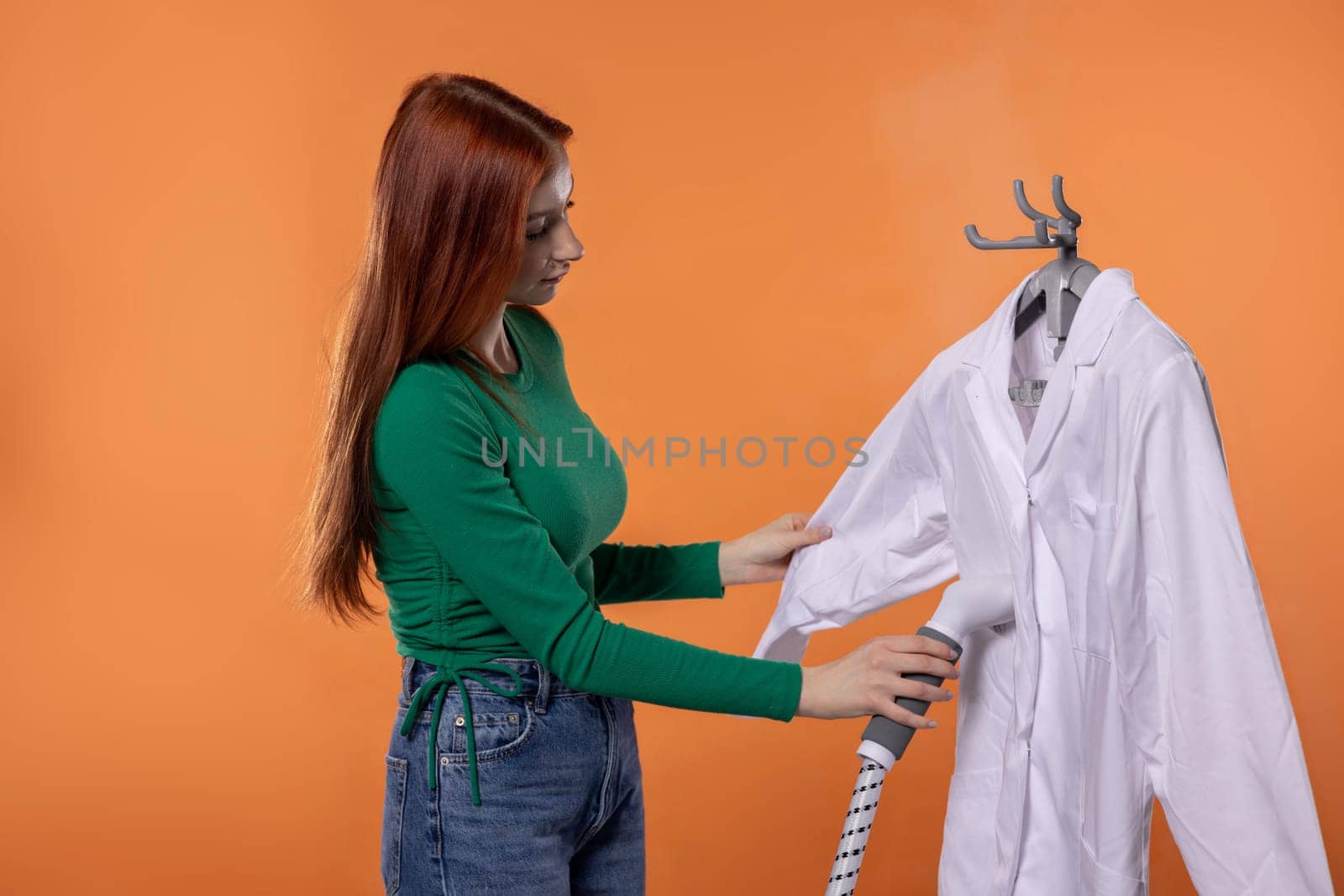 A woman carefully inspects a white shirt hanging on a hanger, preparing it for steaming against a warm orange backdrop