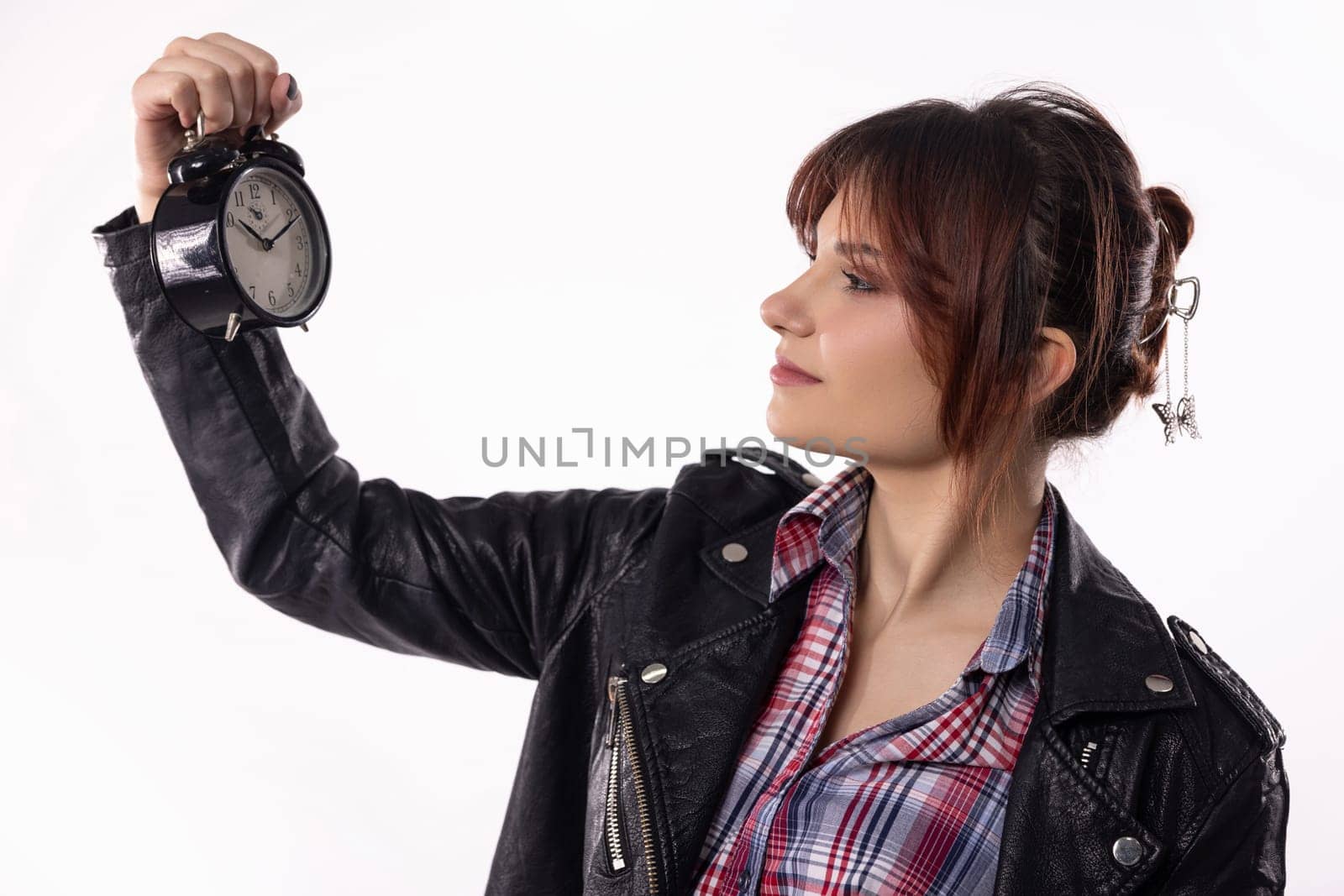 A young woman in a leather jacket and plaid shirt thoughtfully examines an alarm clock, reflecting on time