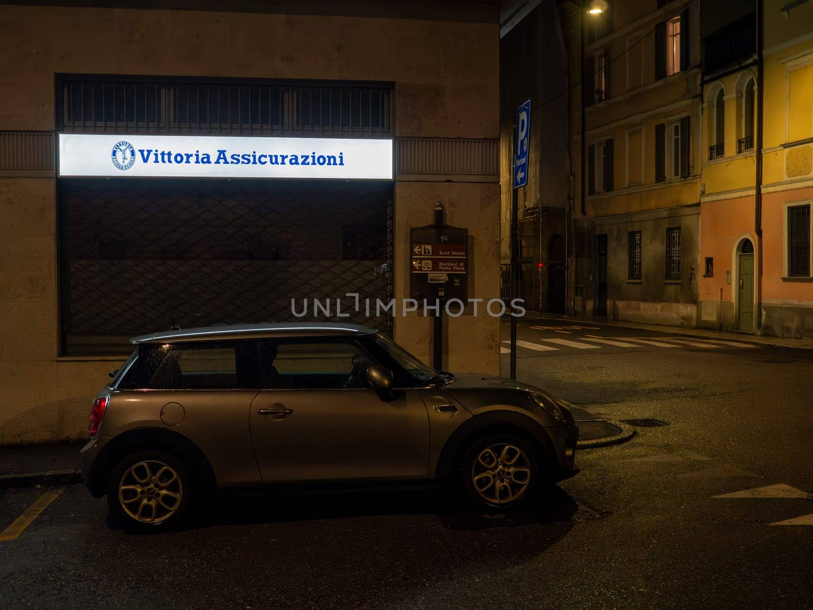 Cremona, Lombardy, Italy October 15th 2024 Parked car sitting on street in front of closed insurance company with illuminated sign at night