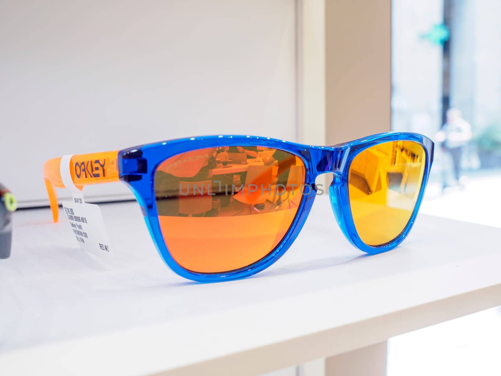Blue sunglasses with orange lenses are resting on a white shelf inside a store, reflecting the interior lights and the street outside