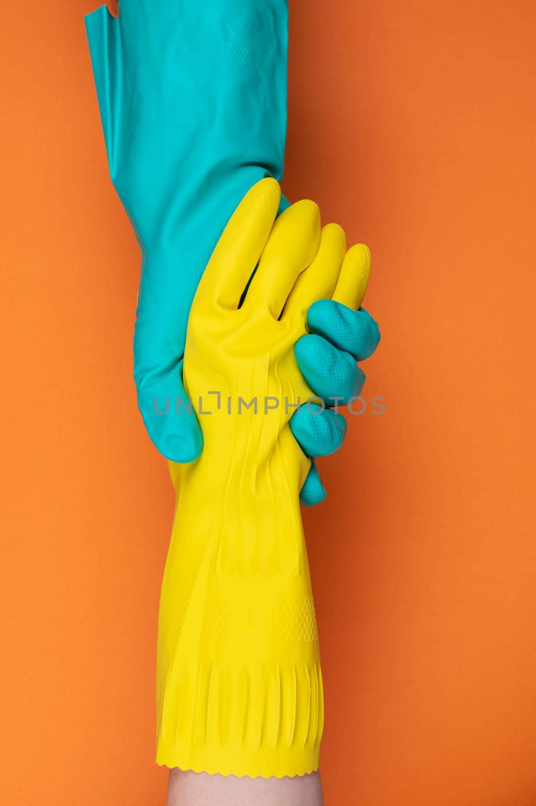 Two hands wearing green and yellow rubber gloves hold each other against a vibrant orange background, symbolizing teamwork and support.