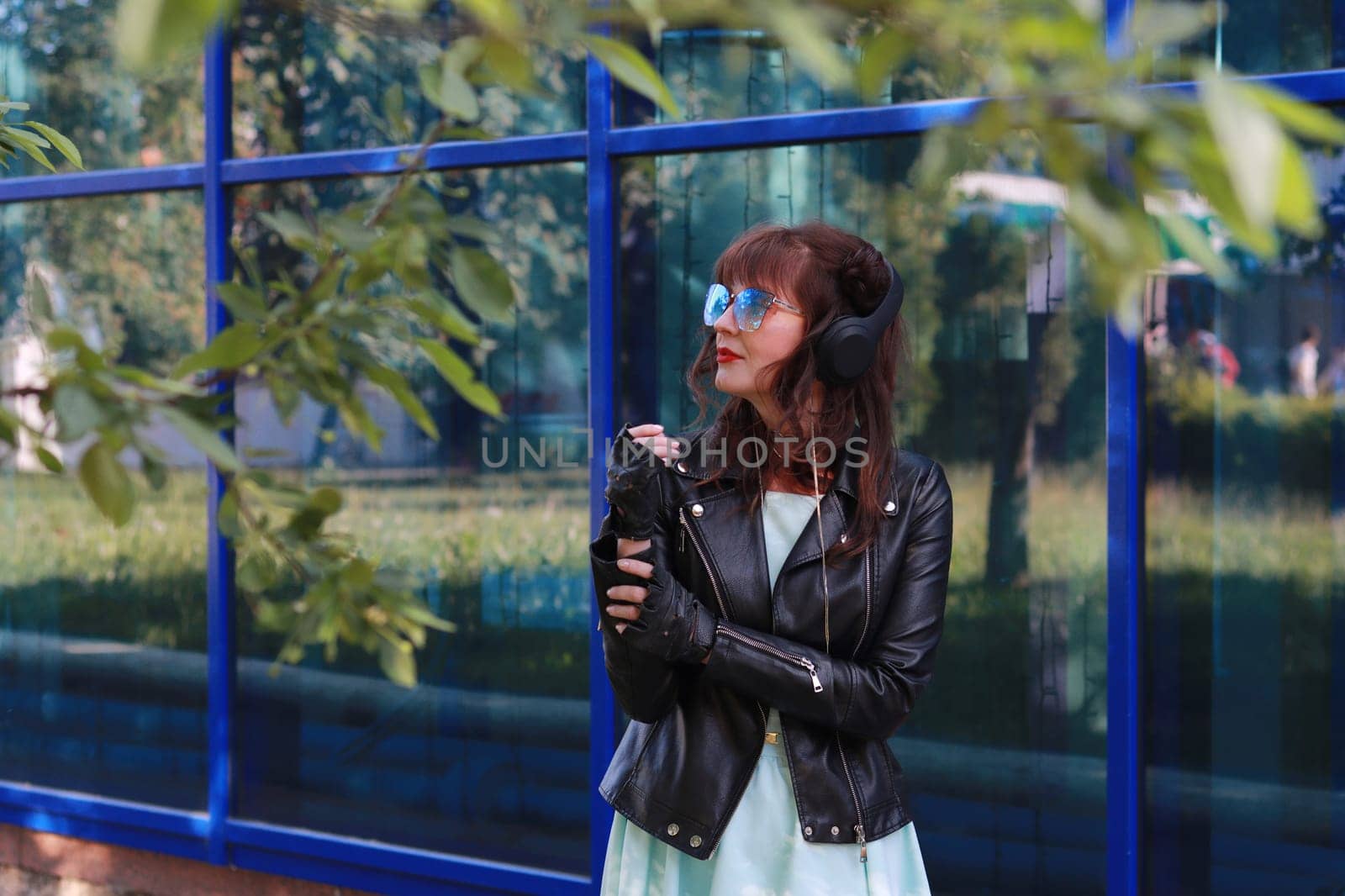 Image of a girl in a blue dress, black leather jacket, wearing blue glasses looking away against a blue glass wall.