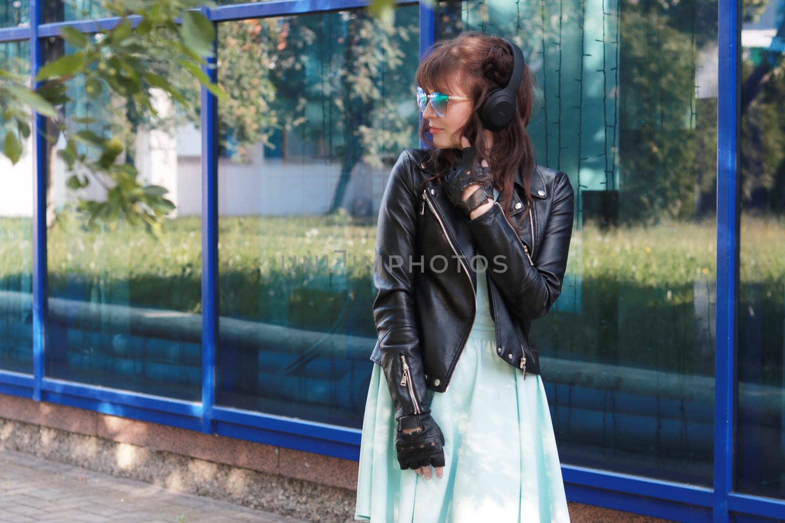 Image of a girl in a blue dress, black leather jacket, wearing blue glasses looking away against a blue glass wall. Adult Girl looking away.
