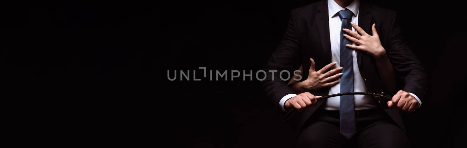 businessman man in a dominant suit with a leather whip is sitting in a chair. Submissive girl puts her arms around him. BDSM concept. Panoramic wide horizontal photo for banner head cover
